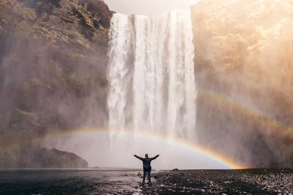Persoon staat met gespreide armen voor een waterval met een regenboog in de nevel